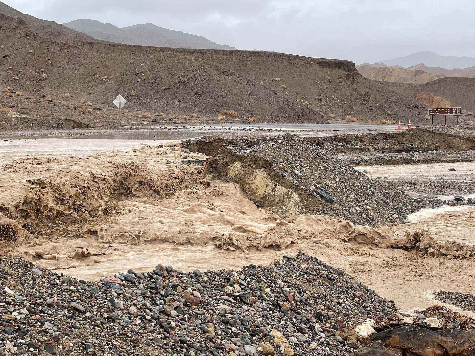 Muddy floodwater flowing through a desert landscape across a paved road causing undercutting on the downhill side. Flash Flooding over Badwater Road near the junction with CA 190. Keywords: Death Valley National Park; Hurricane Hilary; Flash Flooding. Spring flash floods compared with summer heat for unusual roadside stops in 2027