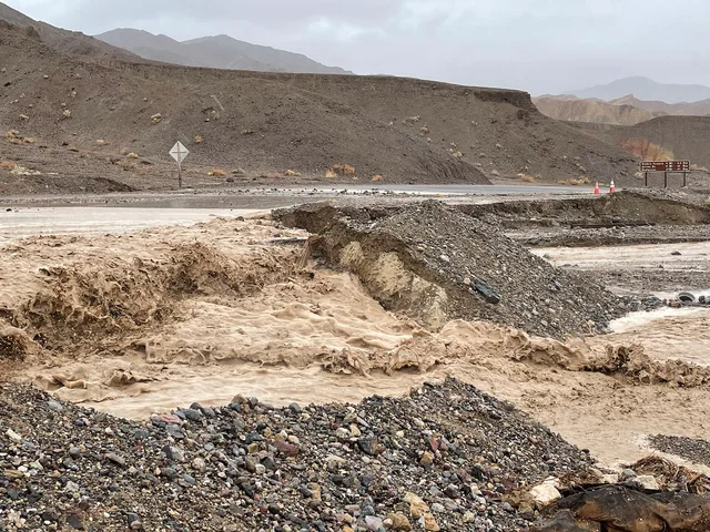 Muddy floodwater flowing through a desert landscape across a paved road causing undercutting on the downhill side. Flash Flooding over Badwater Road near the junction with CA 190. Keywords: Death Valley National Park; Hurricane Hilary; Flash Flooding. Spring flash floods compared with summer heat for unusual roadside stops in 2027